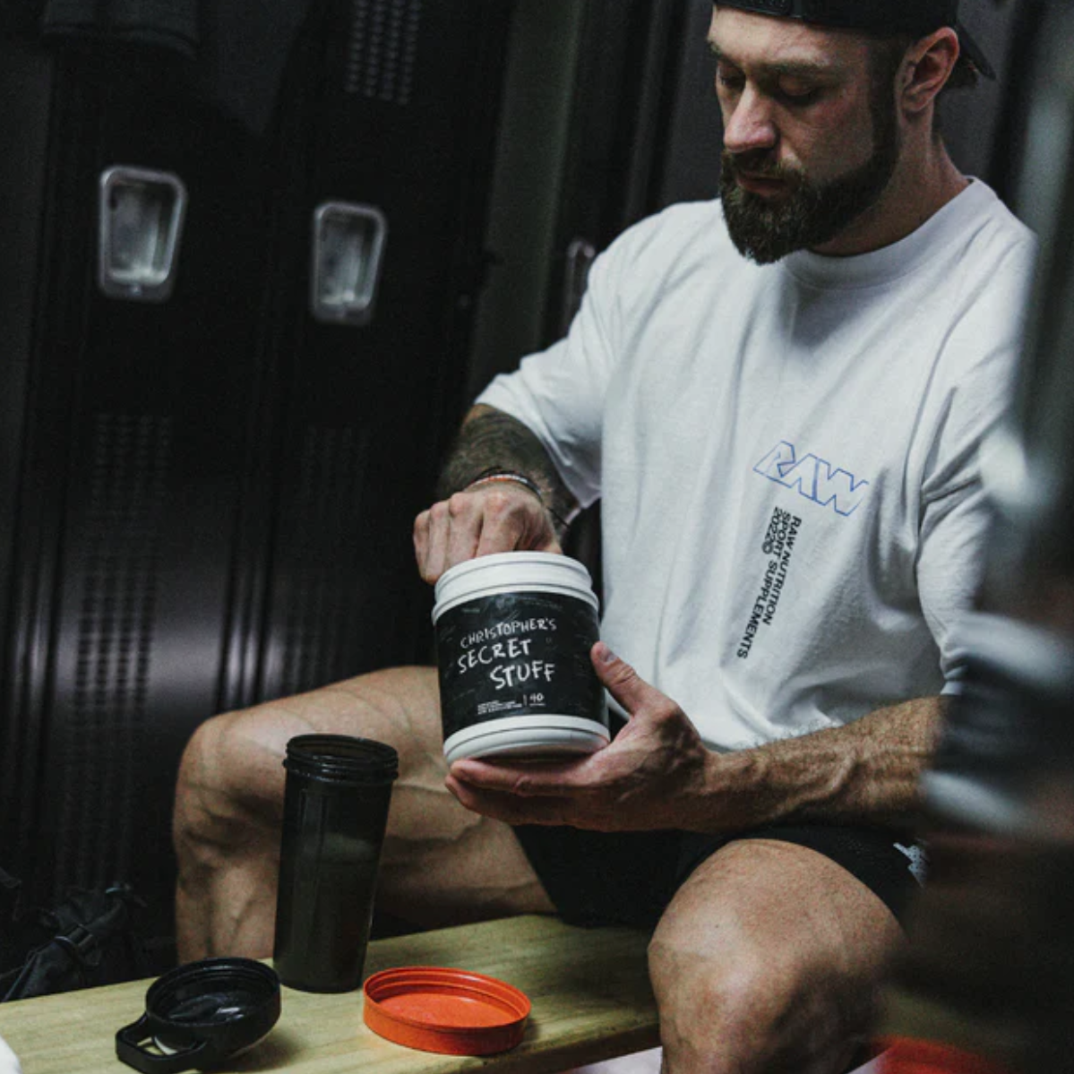 Chris Bumstead is sitting on a bench in a gym locker room, wearing a white oversized t-shirt with "RAW" branding and a black backwards cap. He is holding a container of his CBUM Thavage Pre-Workout labeled "Christopher’s Secret Stuff" and is scooping the pre-workout powder into a black shaker cup. The gym lockers in the background emphasize the locker room setting, while his focused expression indicates he is preparing his pre-workout drink.