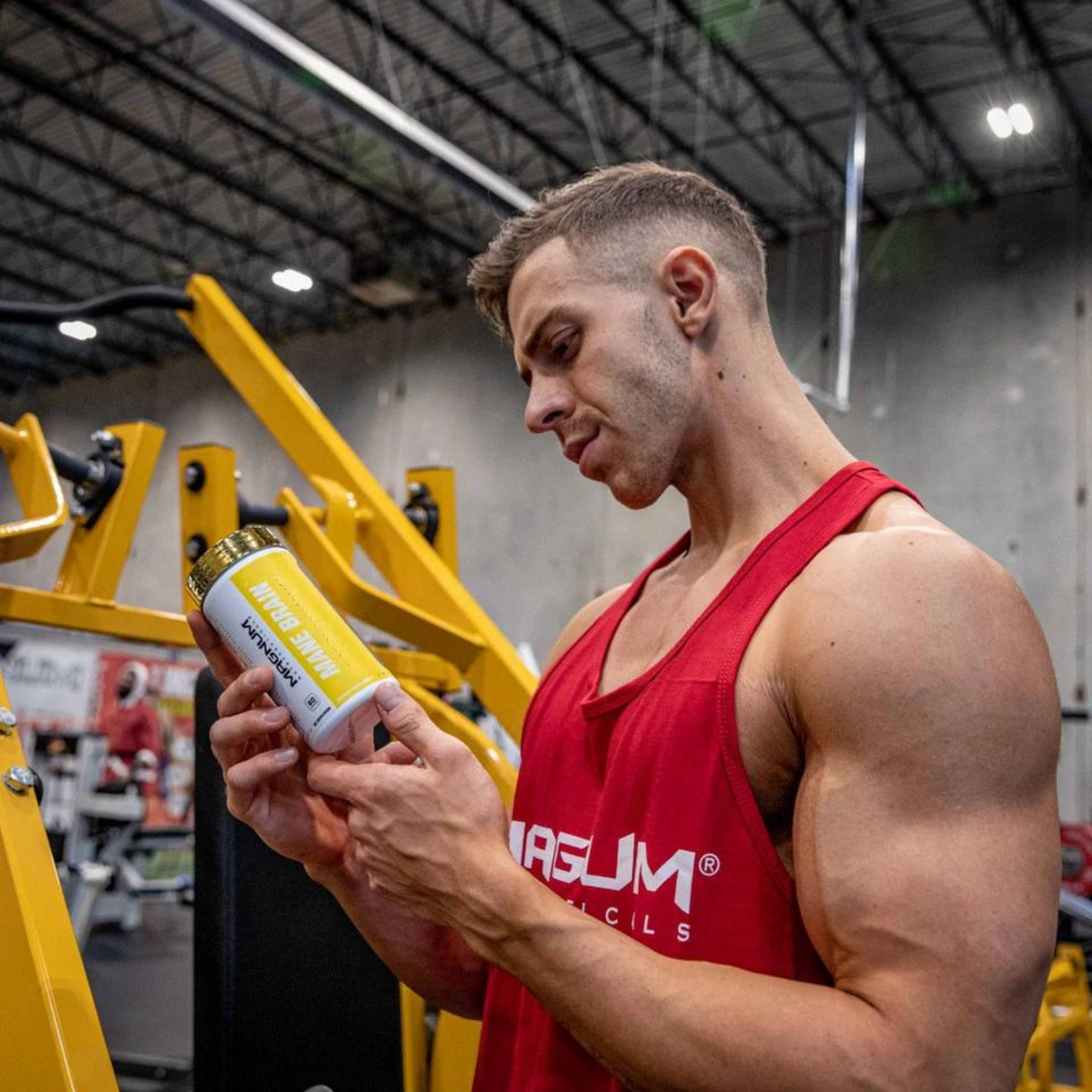 A muscular man in a gym setting, wearing a red Magnum Nutraceuticals tank top, examines a bottle of Magnum Nutraceuticals Mane Brain