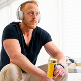 A man wearing headphones sits indoors, holding a container of Magnum Nutraceuticals Mane Brain.