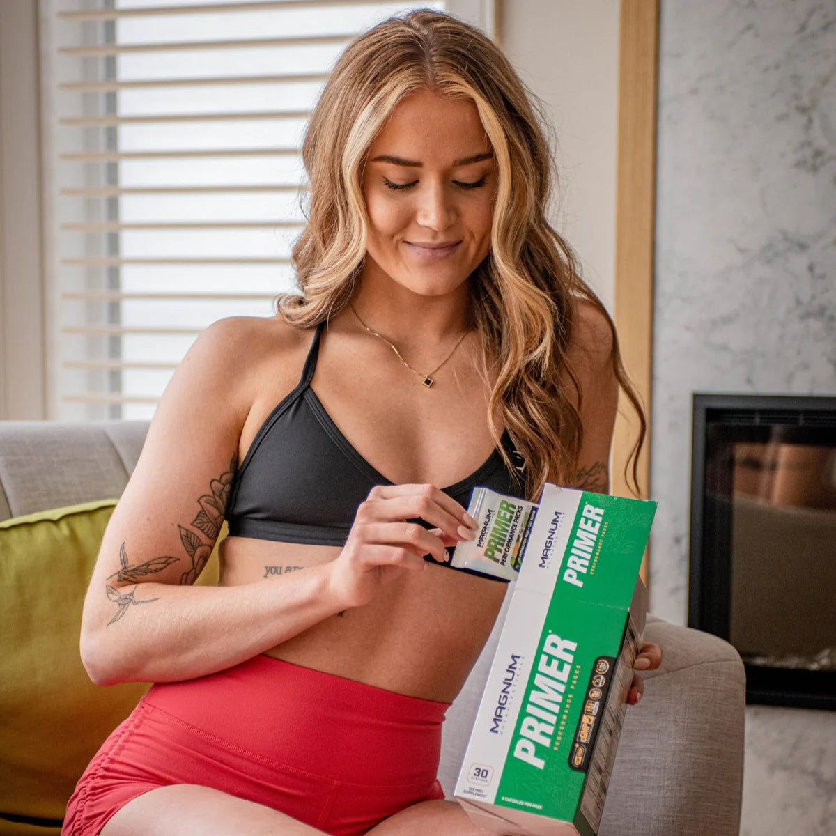 A woman sitting on a couch in a casual indoor setting, wearing a black sports bra and red shorts. She is holding a box of Magnum Nutraceuticals Primer Multivitamin and pulling out a packet from the box. She is smiling softly and appears to be relaxed and focused on the product.