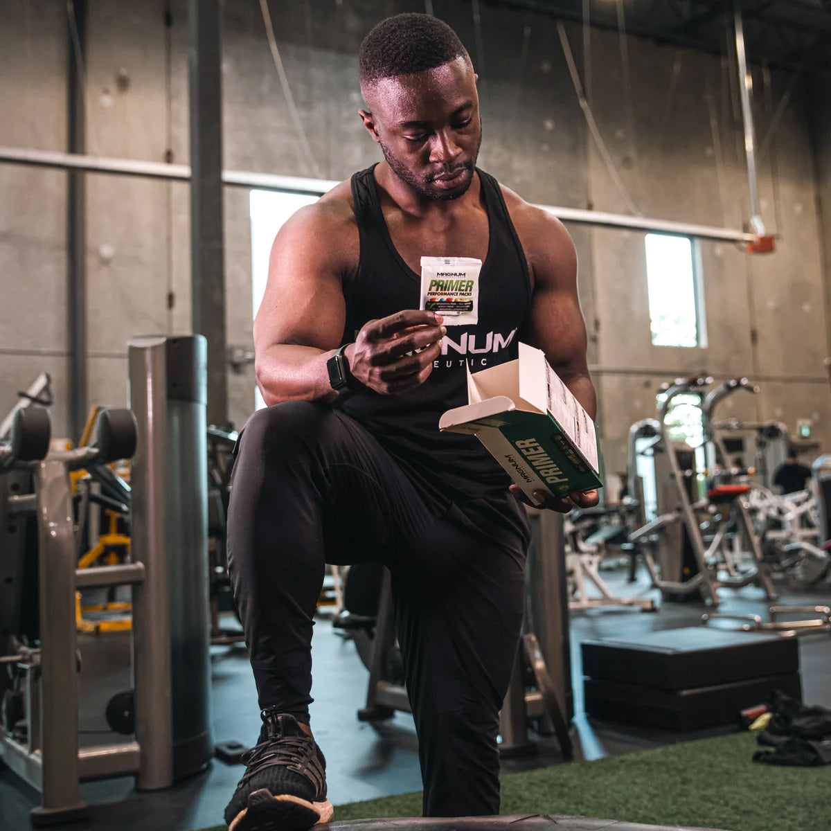 A man in a gym setting, dressed in black athletic wear, holding a box of Magnum Nutraceuticals Primer Multivitamin. He is standing with one foot on a gym bench, looking down at a packet he is holding, with an expression of concentration.