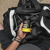 A person is seated with a black duffel bag on their lap, holding a bottle of Opti-Men Multivitamin by Optimum Nutrition. The multivitamin bottle, labeled with a black and yellow design, is prominently displayed in the hand.